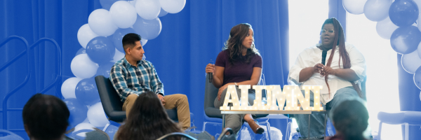 Alumni panelists (two women and one man) speaking to students. Small marquee letters spell out "Alumni" on the table in front of them.