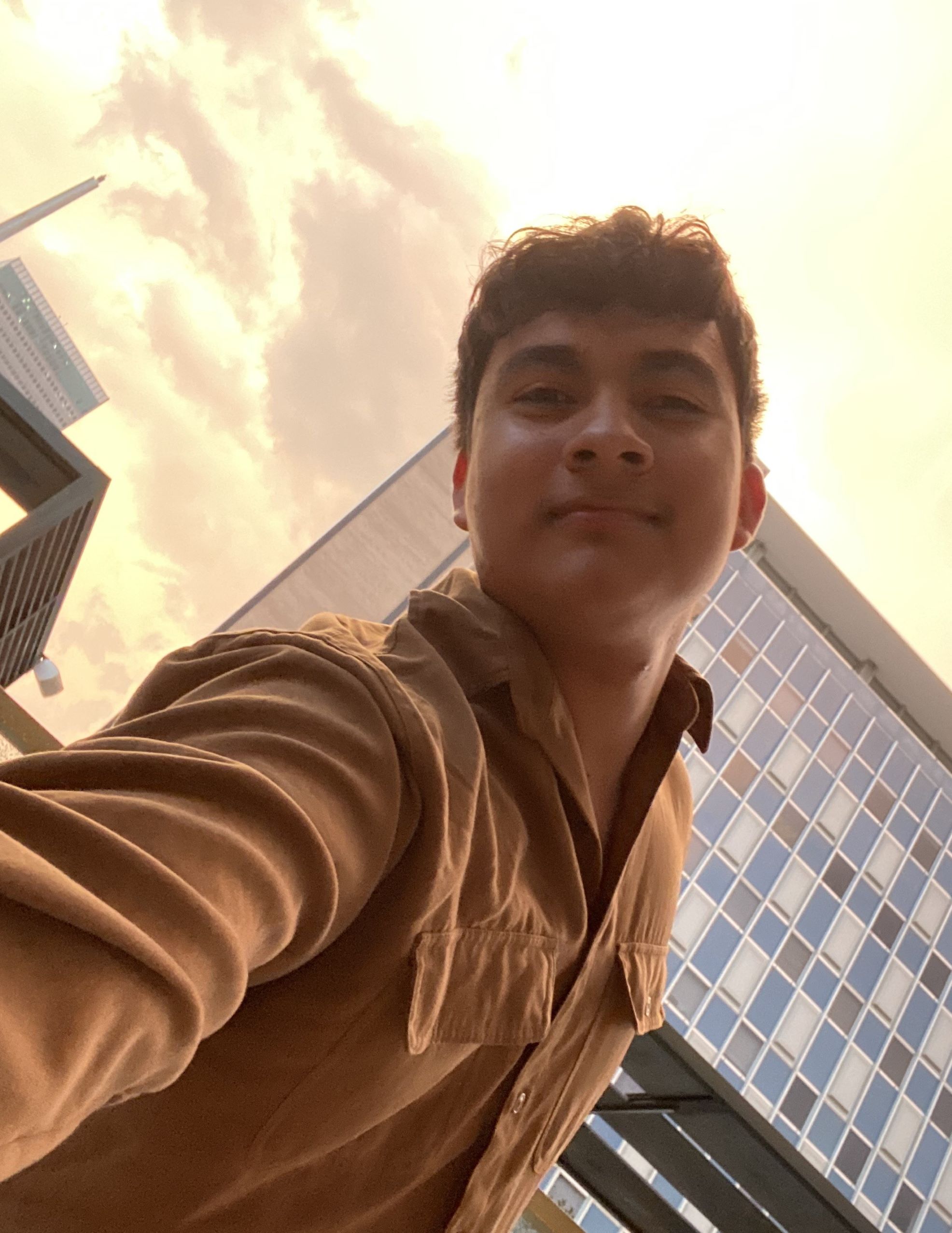 young male in light brown shirt staring down at camera with buildings and a sunset sky behind him