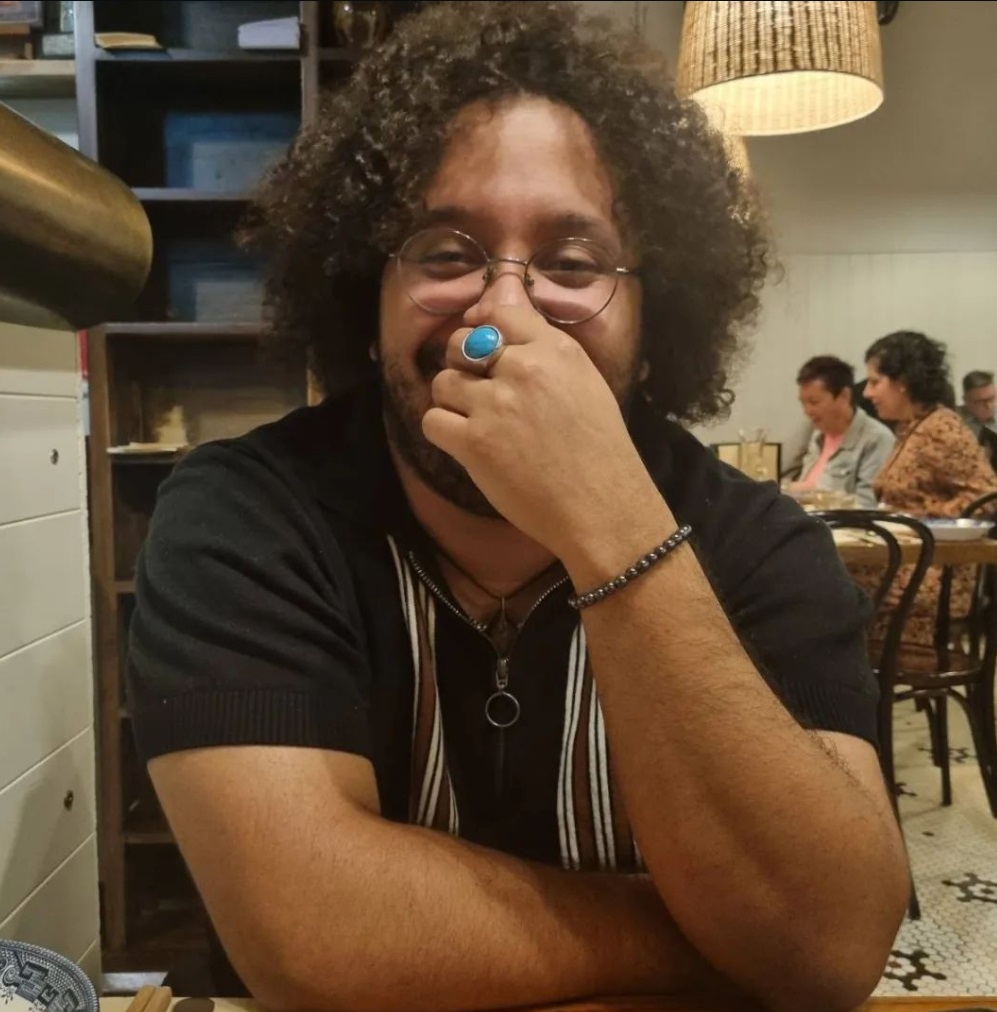 young man with curly hair in a black shirt with whit and brown stripe paneling. person also has a blue ring and dark bracelot